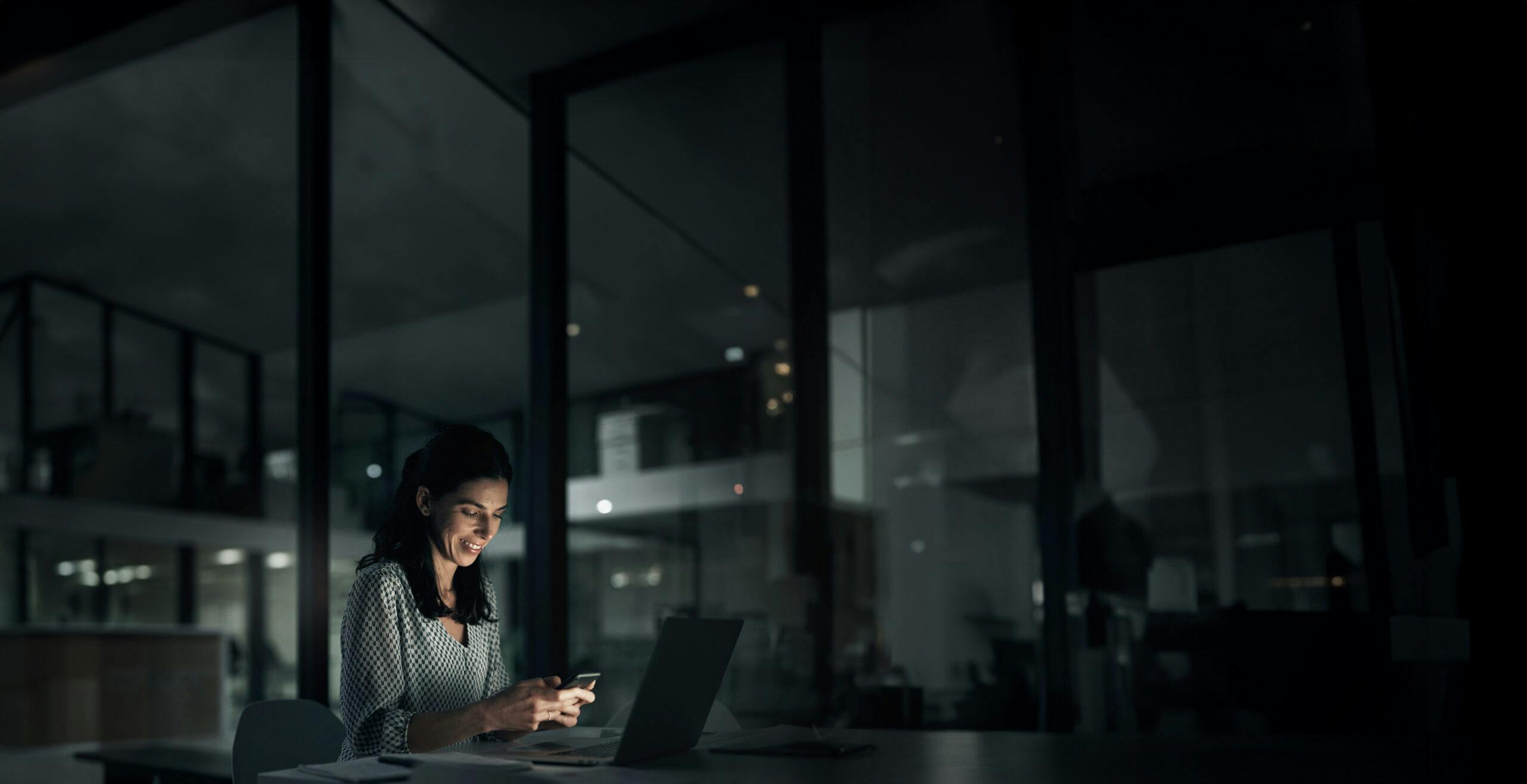 Night, woman working and phone with a smile in a office with planning and digital research. Startup, working and laptop with female person in a company with text reading and mobile networking in dark
